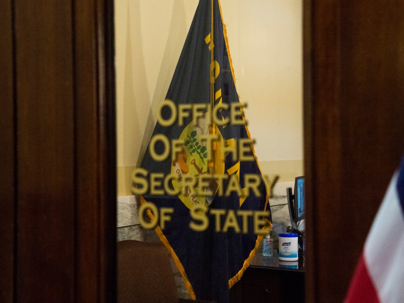 A partially obscured view through an open doorway into an office. "Office of the Secretary of State" is prominently displayed. The edge of an American flag can be seen on the right side of the image.