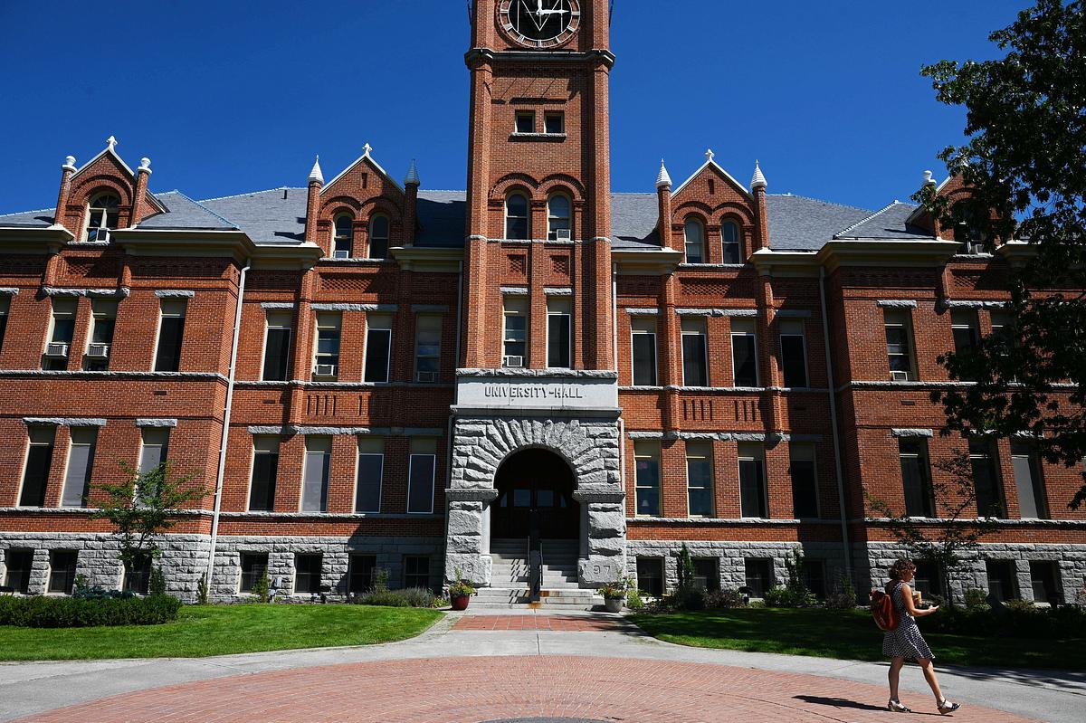 A red-brick University Hall building with a central clock tower and arched stone entrance, set against a clear blue sky, with a person walking along a paved path in the foreground.