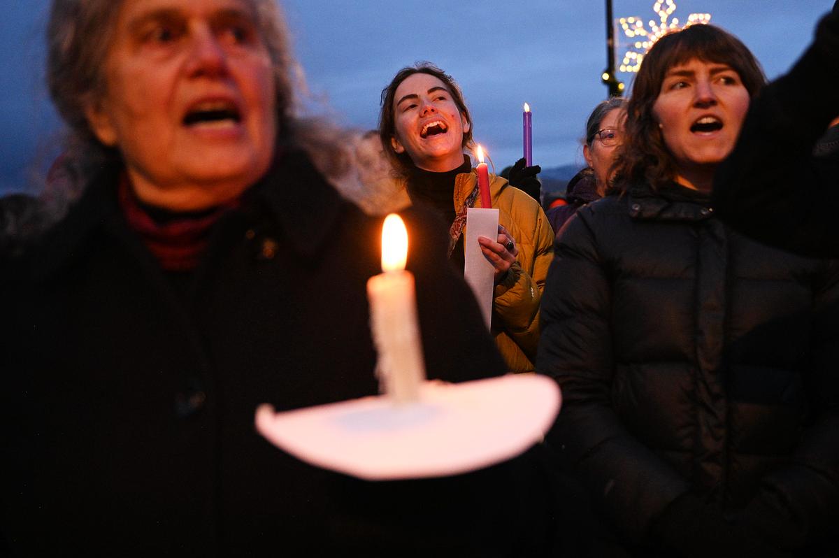 People holding lit candles at an outdoor vigil at dusk, with several faces visible in winter coats and candle flames glowing in the foreground.