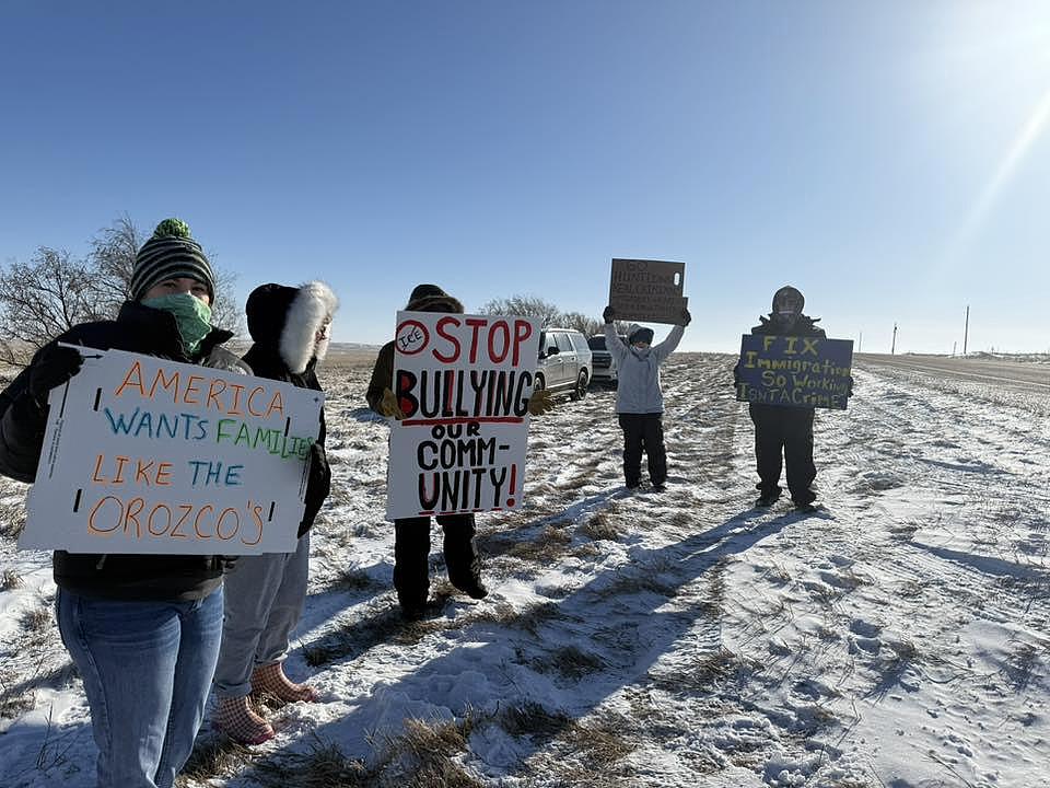 People stand along a snowy roadside holding protest signs supporting immigrant families and community unity.